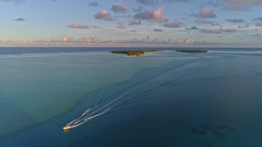 Aerial view of a boat cruising on a beautiful turquoise lagoon with tropical islands on the horizon at sunrise