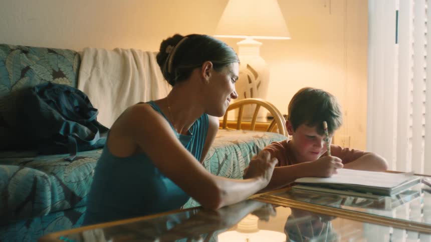 A smiling mother watches and helps her young son as he concentrates on drawing in a notebook at a coffee table in a warmly lit room