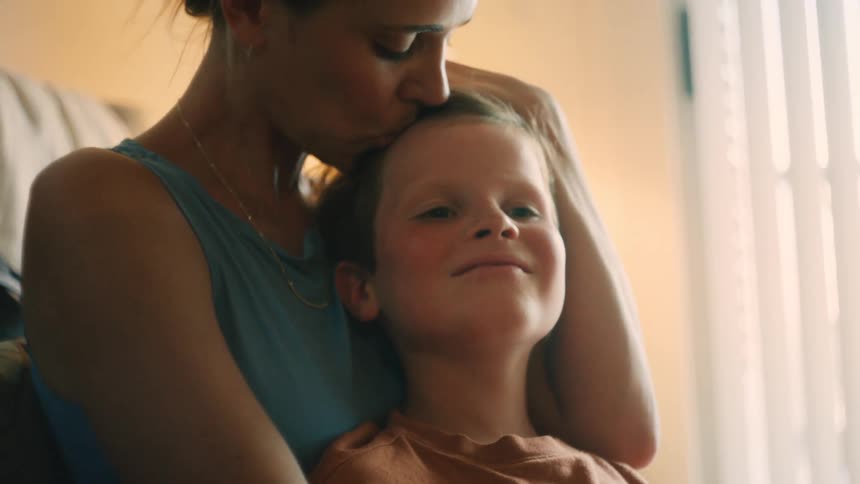 In a softly lit room, a mother lovingly caresses her son's hair and kisses his forehead in a heartwarming embrace