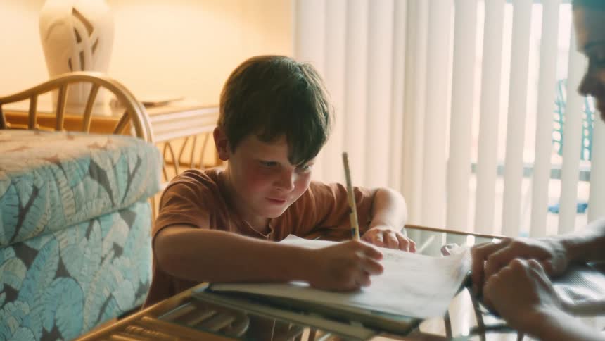 A mother smiles as she helps her playful son with his homework at a sunlit table in their home