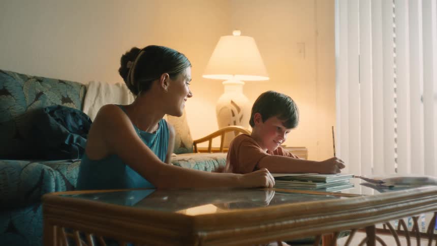 A caring mother smiles and encourages her young son as he works on his homework at the coffee table in their sunlit living room