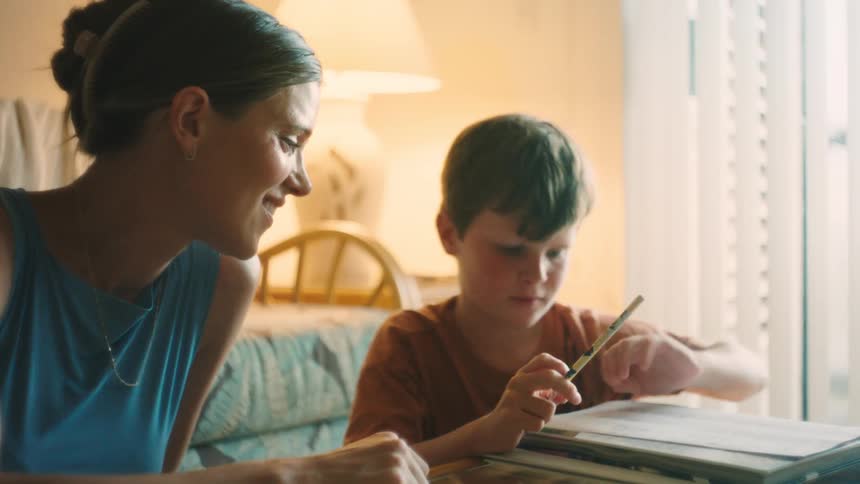 A smiling mother patiently helps her young son with homework at a table in their warm, sunlit living room