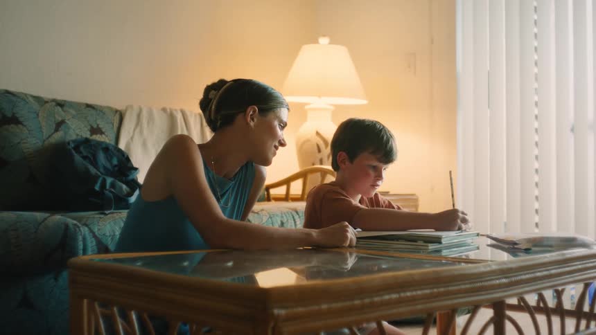 A proud mother lovingly watches her young son as he draws in a sketchbook at their cozy living room coffee table