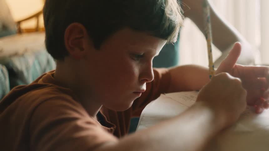 A young boy concentrates on his homework as his supportive mother sits beside him and points to his paper to help him