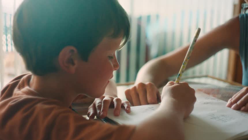 A young boy smiles while doing homework with his mother's help at a sunlit table, then refocuses with concentration
