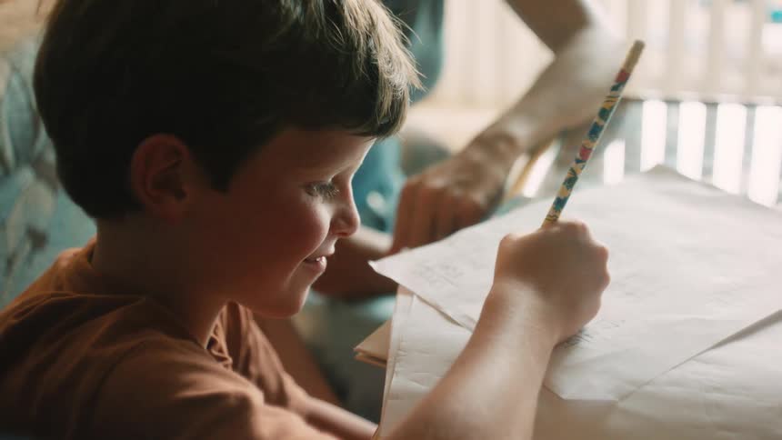 A young boy with freckles focuses intently on his homework, writing with a pencil as his mother helps him at the table in slow motion