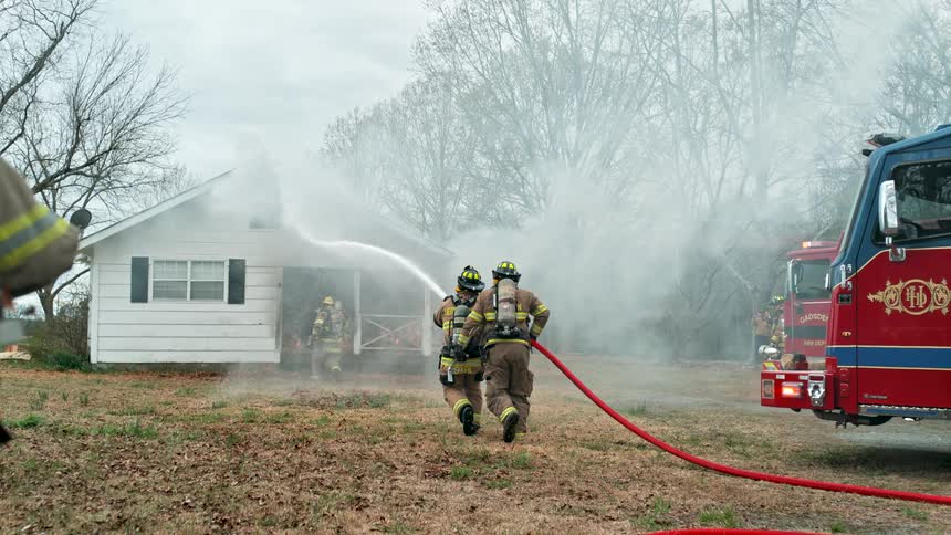 Firefighters in full gear work together to extinguish a house fire with a high-pressure water hose as smoke billows from the building