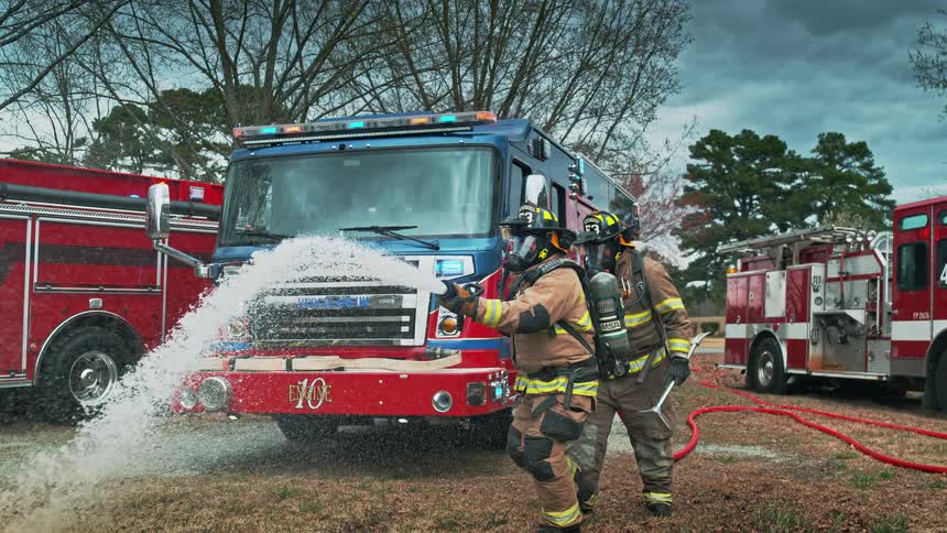 Two firefighters in full gear spray a powerful stream of water from a fire hose in dramatic slow motion with fire trucks behind them