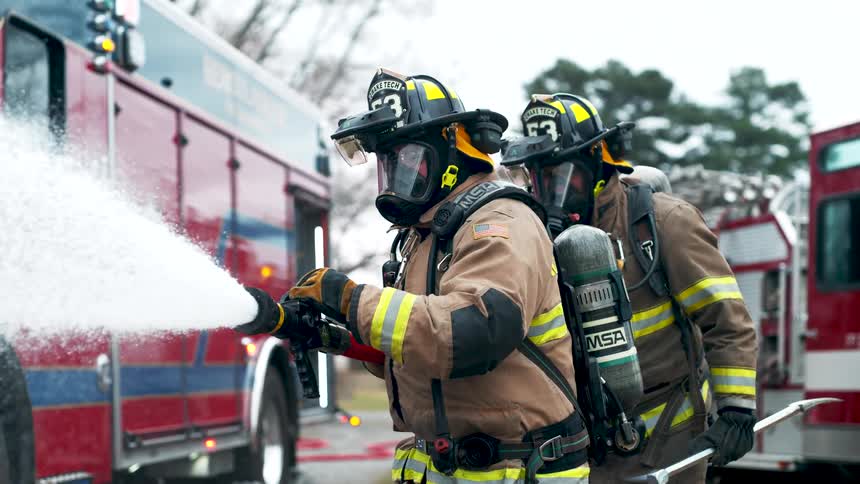 Two firefighters in full gear stand next to a fire truck as one sprays a fire hose in slow motion