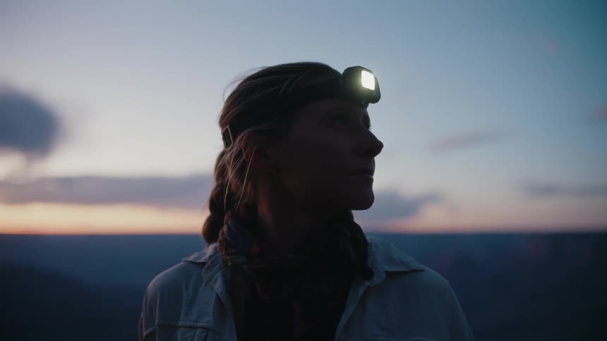 A young woman with braided hair turns on her headlamp as she gazes at the beautiful twilight sky over the mountains