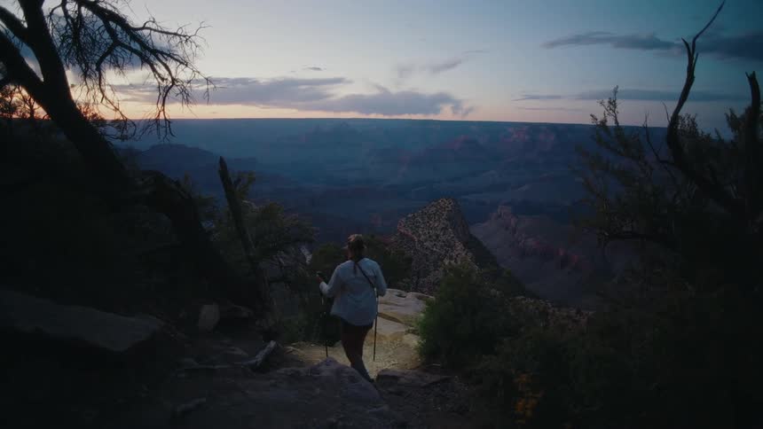 A female hiker with trekking poles walks in slow motion along a trail to a scenic overlook of the Grand Canyon at sunrise