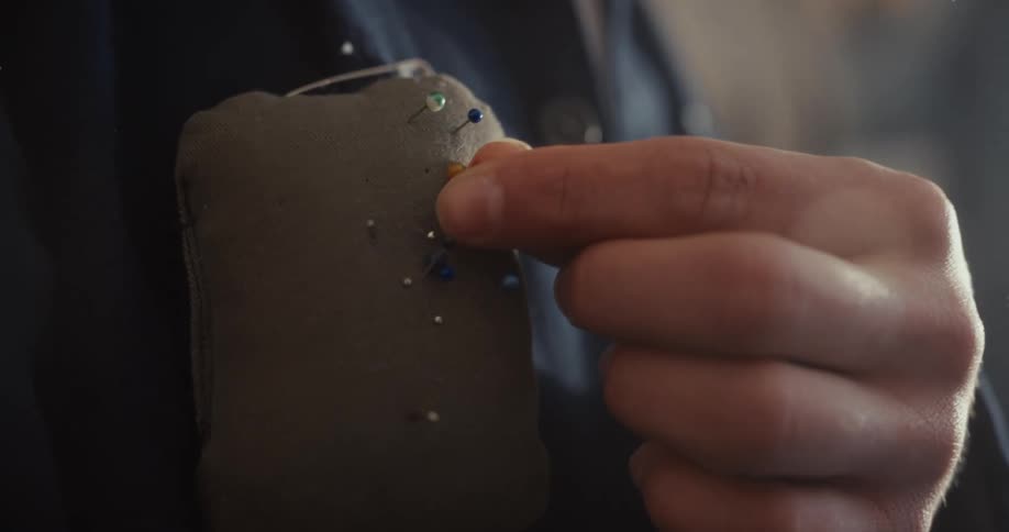 A close-up of a tailor's hand carefully placing a colored pin into a pincushion attached to their jacket in slow motion