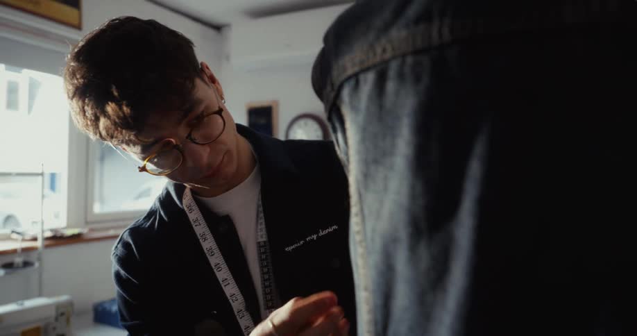 A skilled male tailor carefully pins and adjusts a denim jacket on a mannequin in his sun-drenched atelier