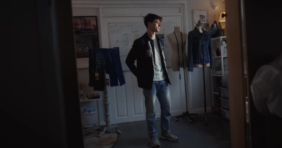 A young male fashion designer stands thoughtfully in his sunlit studio, surrounded by mannequins and sketches