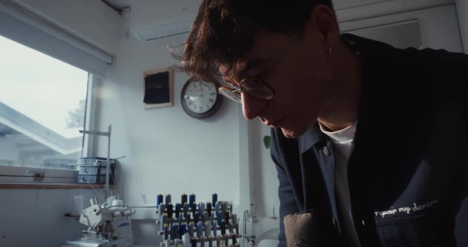 A focused young fashion designer with glasses types on his laptop in a sunlit sewing workshop