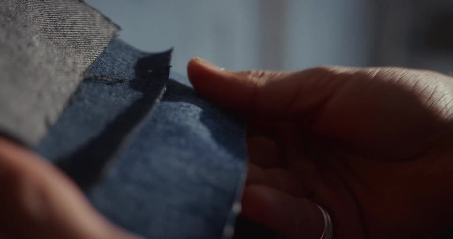A close-up of a designer's hands examining and feeling the texture of various blue denim fabric swatches in slow motion