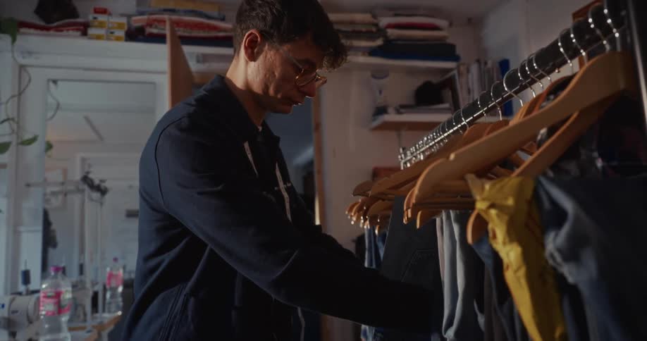 A male fashion designer carefully inspects and hangs a pair of jeans on a clothing rack in his workshop