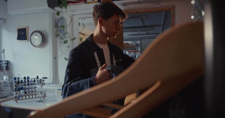 A young male tailor carefully examines a denim garment in his workshop