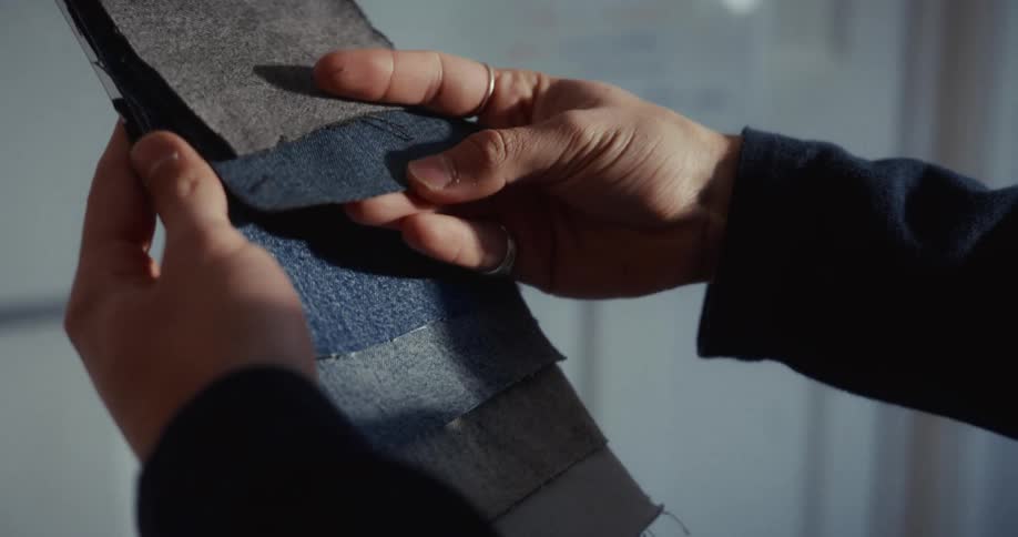 A designer's hands carefully feel and select from a collection of blue and grey denim fabric swatches in a sunlit workshop