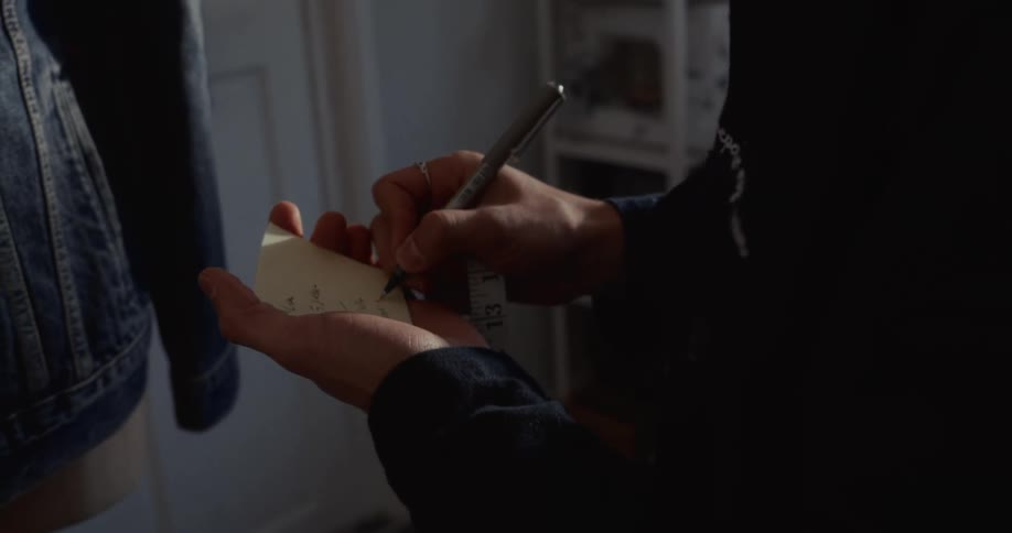 A focused tailor in a dimly lit workshop carefully jots down notes and measurements on a small pad