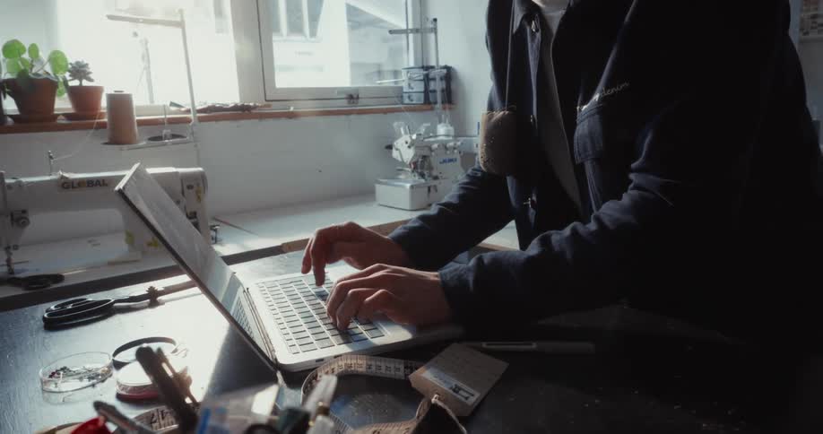 A focused male fashion designer types on a laptop at a workbench in his creative, sunlit workshop