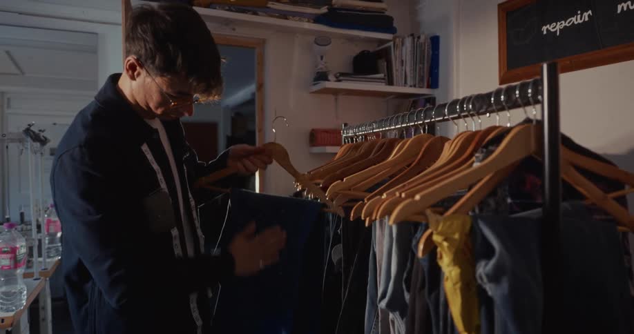 A meticulous male tailor in his workshop carefully inspects a pair of blue jeans before hanging them on a clothing rack