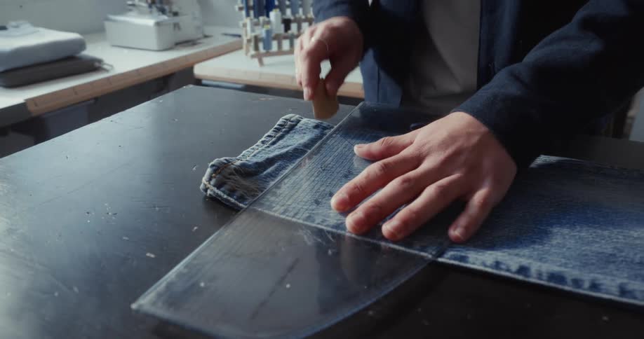 A craftsman uses a template and chalk to carefully mark a cutting line on a pair of blue jeans in a workshop