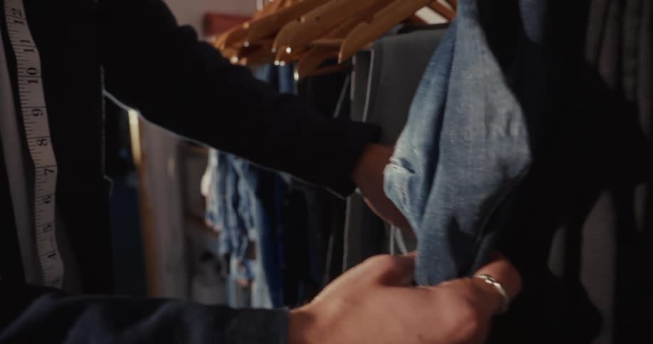 A tailor carefully inspects a collection of denim jeans hanging on a clothing rack in his design studio