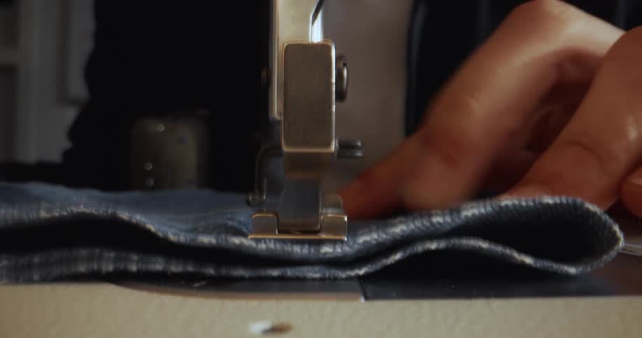 Slow-motion, front-facing close-up of a sewing machine's needle and presser foot stitching blue denim fabric