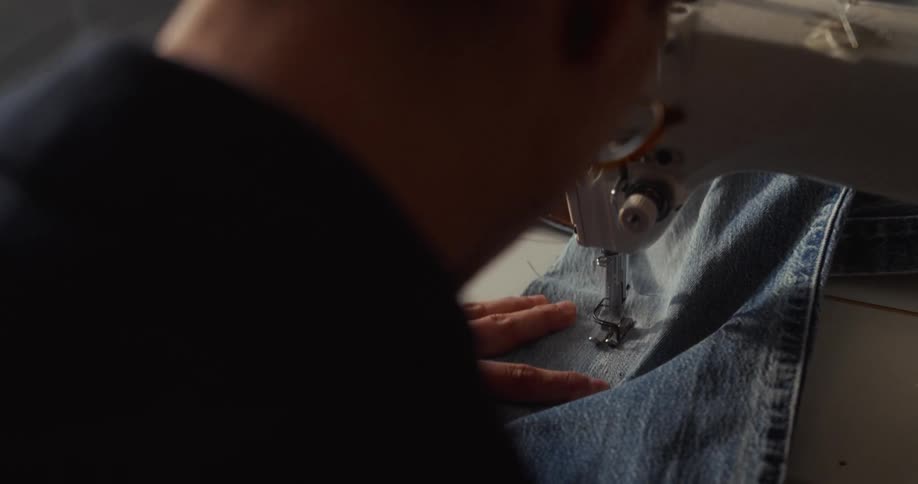 An over-the-shoulder view shows a person carefully guiding denim fabric through a sewing machine