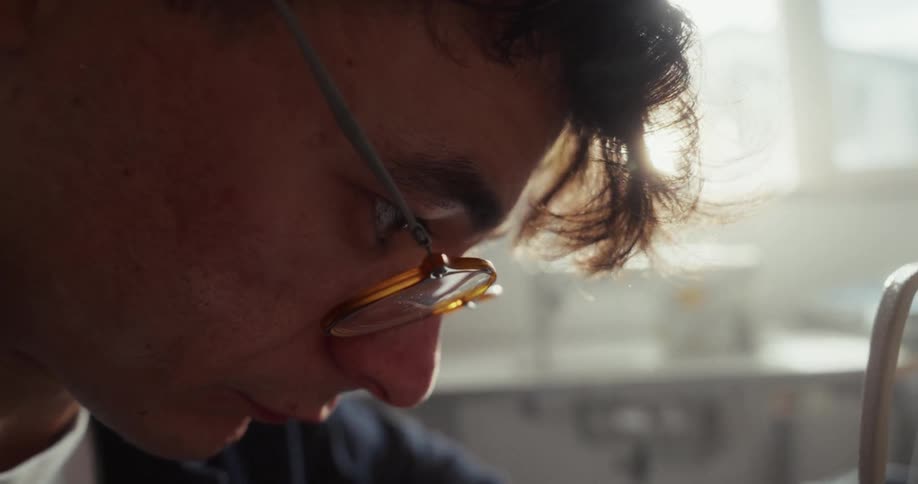 A cinematic close-up of a young man with glasses, his hair backlit by the sun, as he focuses intently on his work