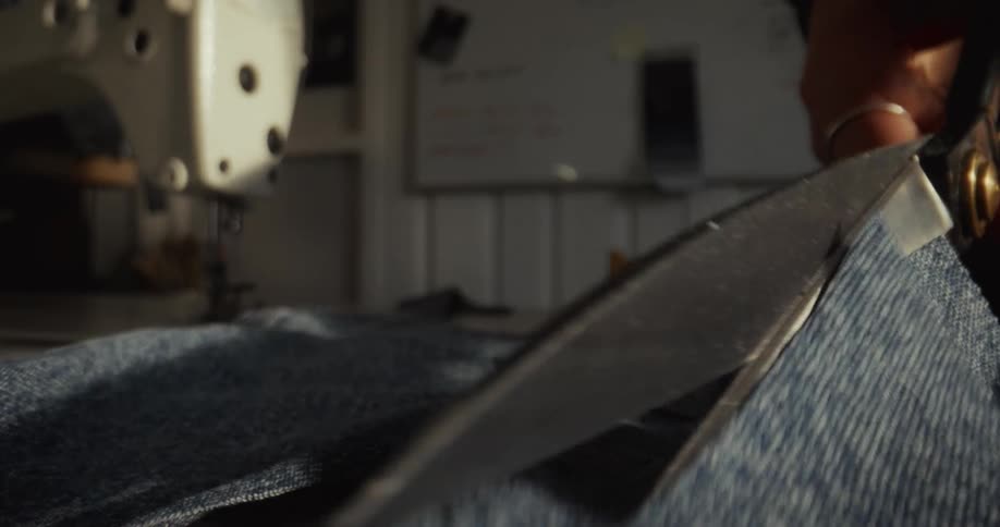 Extreme close-up in slow motion of a tailor cutting through blue denim fabric with large, sharp scissors in a sunlit workshop
