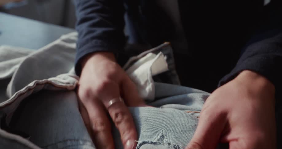 A person carefully inspects a hole in a pair of light-wash denim jeans, examining the fabric closely