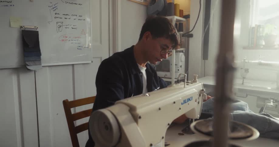 A focused male tailor uses an industrial sewing machine to stitch a piece of denim in his sunlit workshop