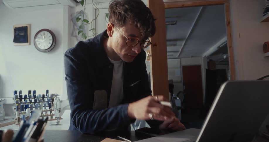 A concentrated young man wearing glasses works diligently on his laptop in a design workshop