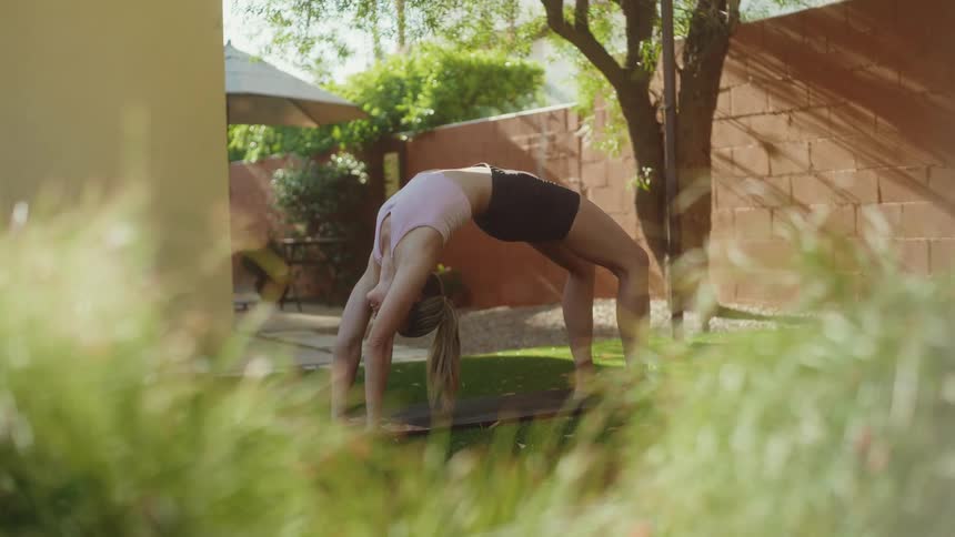 A flexible woman holds a wheel pose on a yoga mat in a sunlit backyard, slowly revealed through green foliage