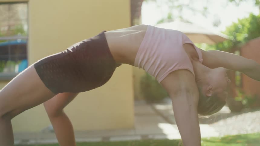 A woman performs a deep backbend yoga pose on a mat in her sunny backyard, with the camera panning across her body