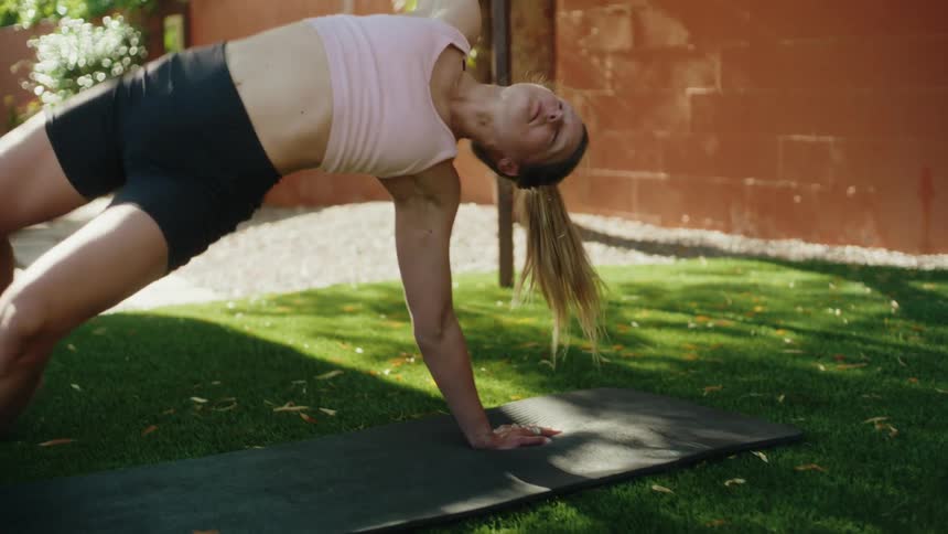 A fit woman gracefully performs a deep backbend yoga pose on a mat in a sunny backyard in slow motion