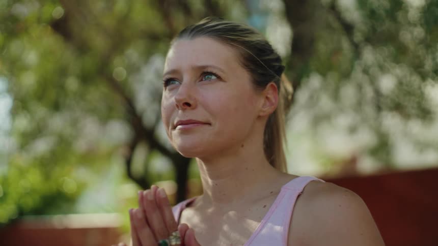A focused woman holds a yoga tree pose with her hands in a prayer position while meditating in a sunny garden