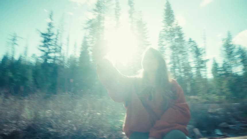 A happy young woman takes a selfie on her phone while resting on a log in a beautiful, sun-drenched forest