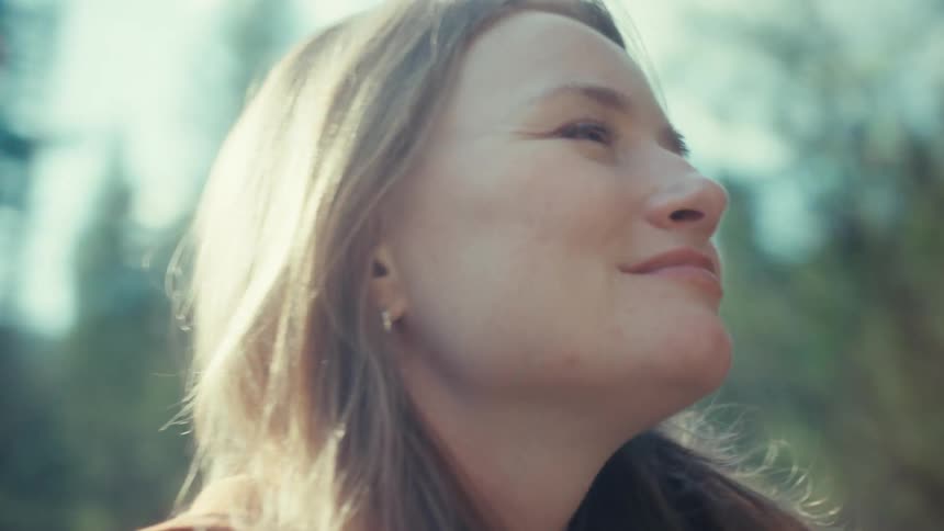 A serene young woman basks in the hazy, dream-like sunlight in a forest, smiling peacefully as she looks up in quiet contemplation