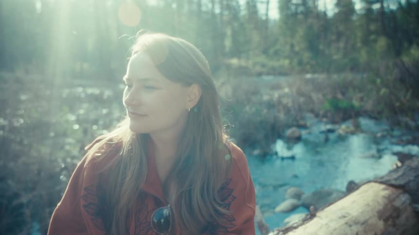 Dreamy slow-motion shot of a happy woman relaxing on a log by a river in a sunlit forest