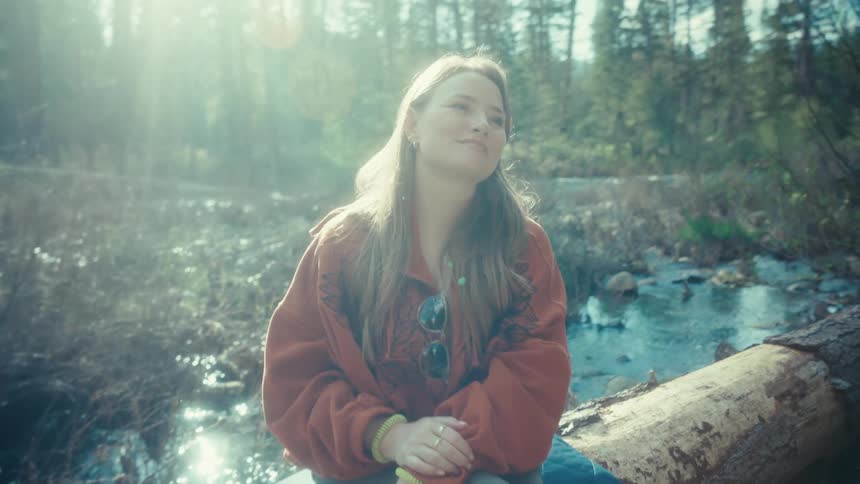 A serene woman in a hoodie enjoys the warm sunshine while sitting on a fallen log next to a tranquil forest stream