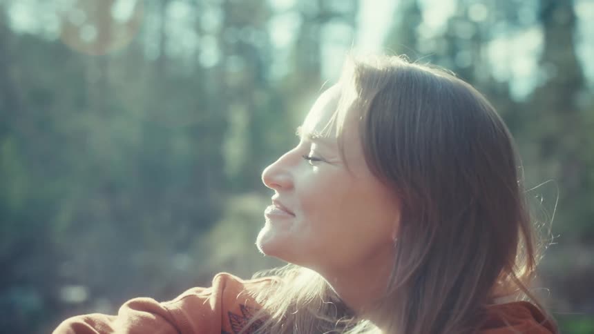 A serene young woman smiles peacefully as she basks in the warm, hazy sunlight while relaxing in a tranquil forest