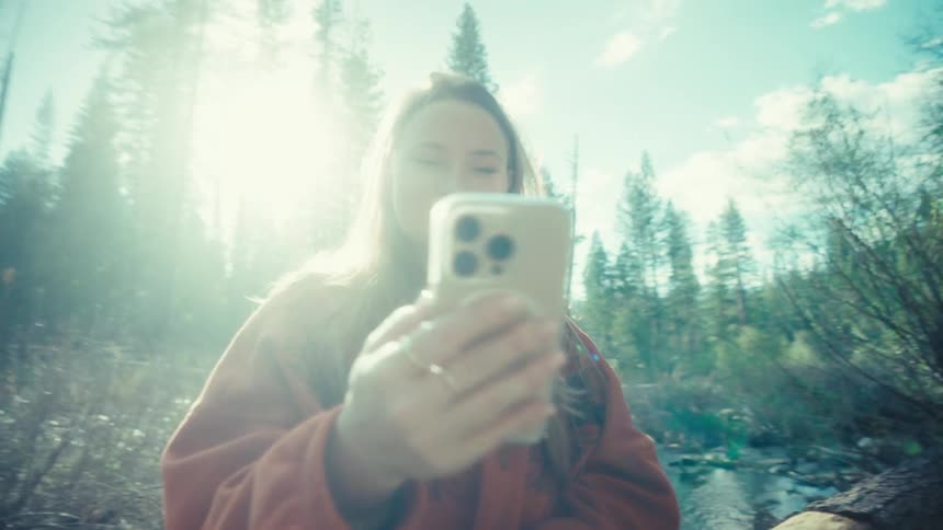 A dreamy, slow-motion push-in shot captures a happy woman smiling while using her smartphone in a sun-drenched forest