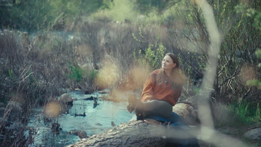 A tranquil woman soaks in the warm sunlight while resting on a log beside a gentle stream in a peaceful forest