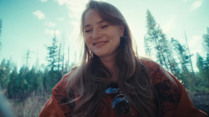 A smiling young woman uses her smartphone while sitting in a beautiful sunlit forest, creating a dreamy lens flare