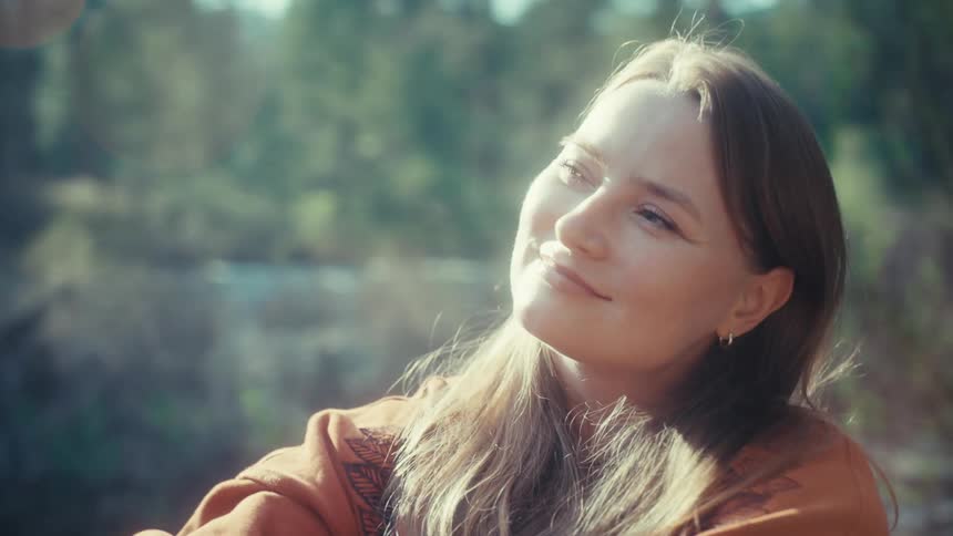 A woman sits peacefully in a sunlit forest, closing her eyes and smiling as she soaks in the warm light
