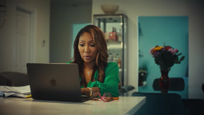 A professional African American woman in a green blouse works from home on her laptop at her modern kitchen island