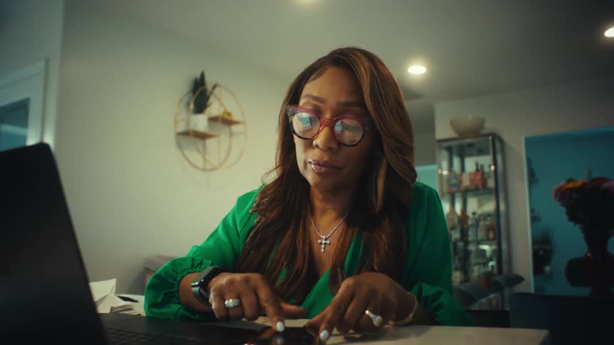 A focused businesswoman in glasses works from home, multitasking on her laptop and smartphone at her kitchen counter